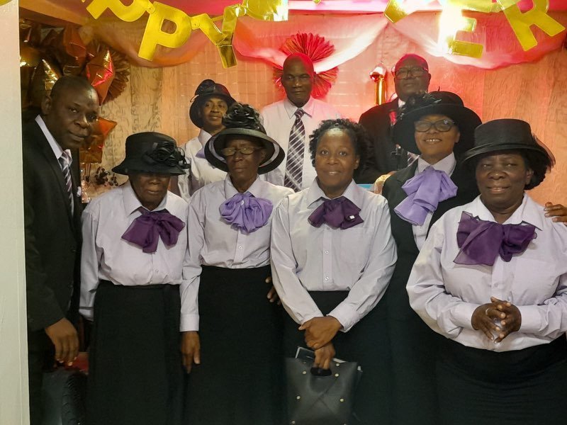 Women of the church in white blouses, purple bows and black hats, standing together with men of the church